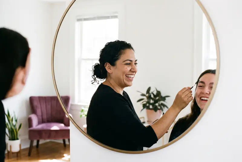 A brow artist laughs with her client in a round mirror while brushing up laminated brows
