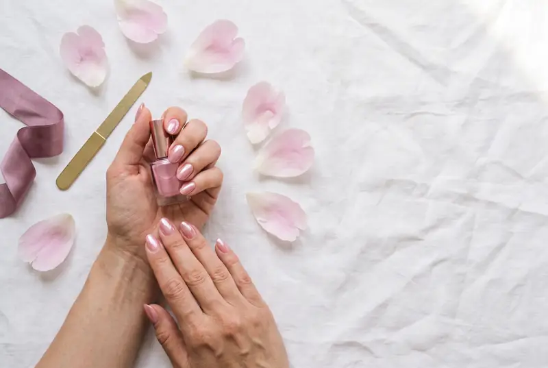 Hands with a sheer pink chrome manicure on white linen with peony petals